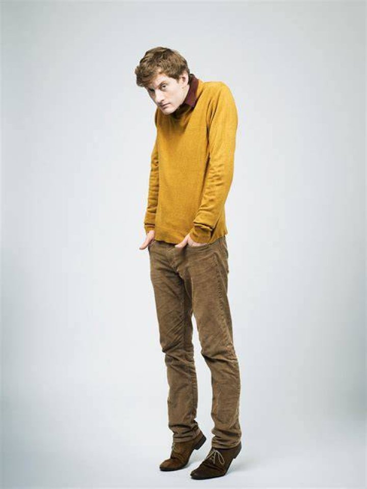 James Acaster is wearing a sweater which has brown, orange, and peach color mixed.